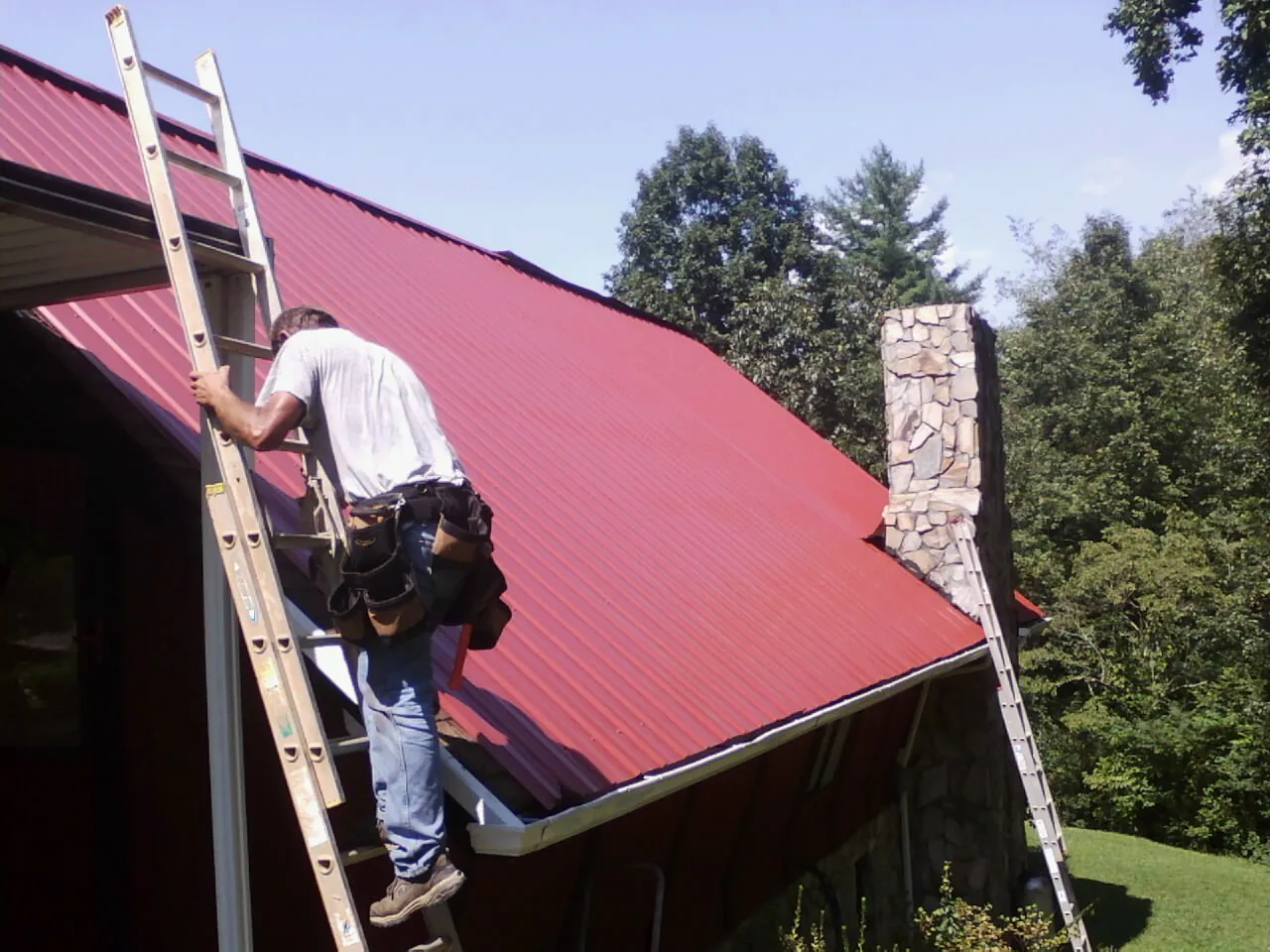 Roofer on a ladder beside a red metal roof and stone chimney — Soffit & Fascia Repair by Roofing Brothers in Independence